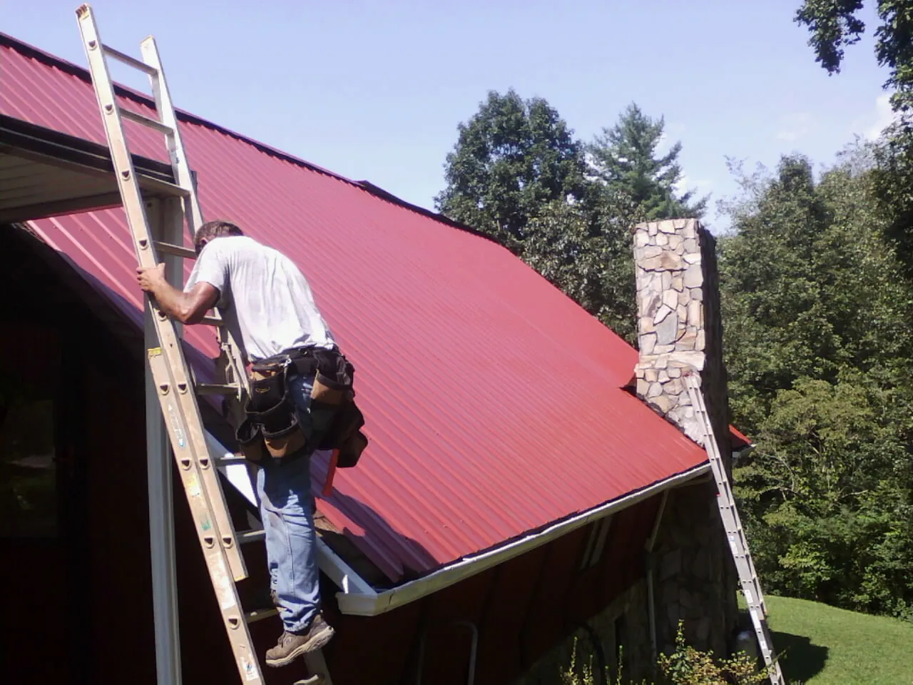 Roofer on a ladder beside a red metal roof and stone chimney — Soffit & Fascia Repair by Roofing Brothers in Elberton